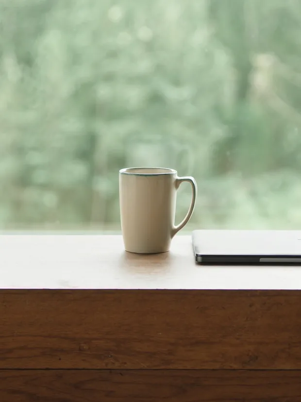 Stoneware cup beside a closed laptop on brown wood, a soft green forest view behind
